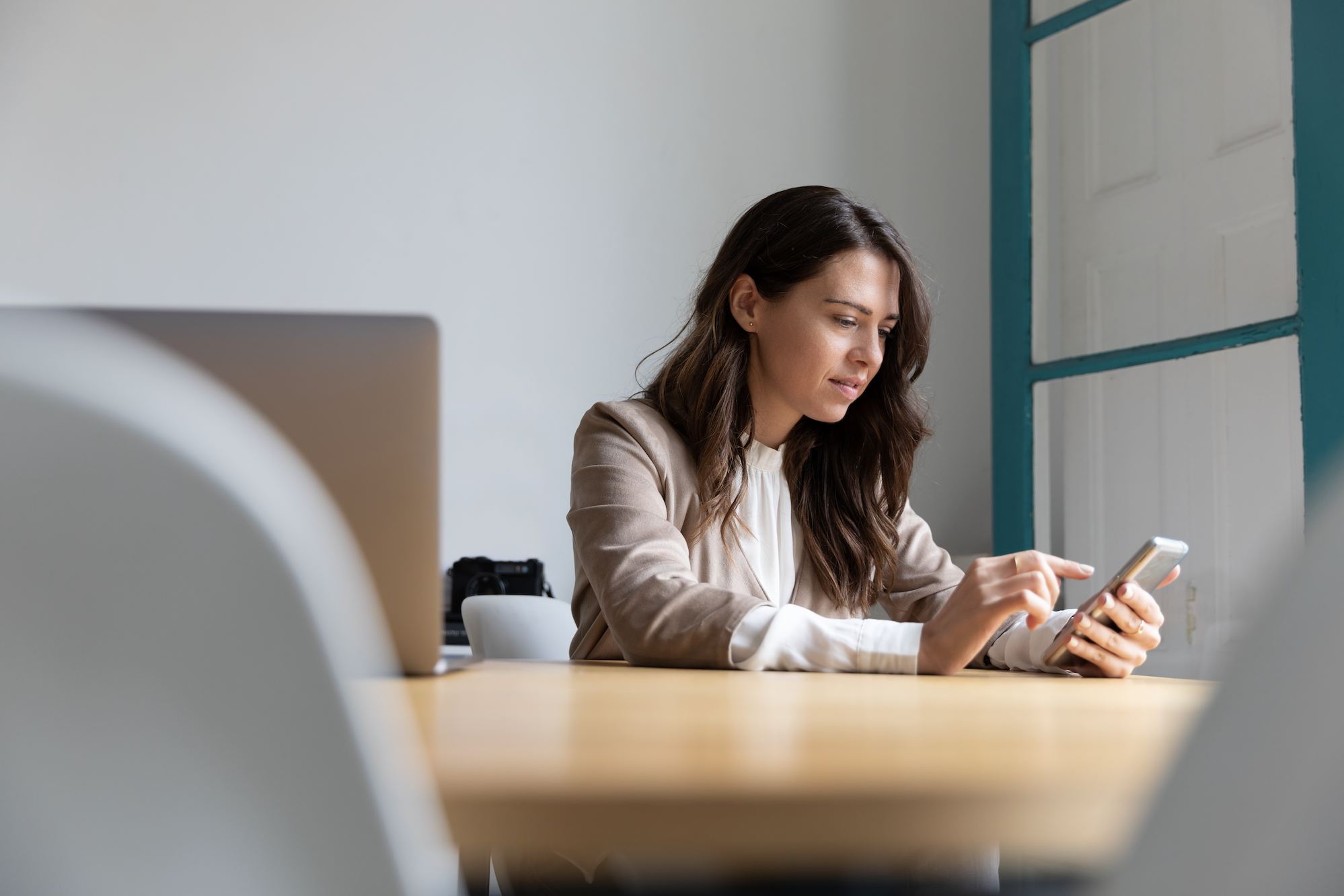 brunette woman sitting at an office desk in a beige blazer using her mobile device